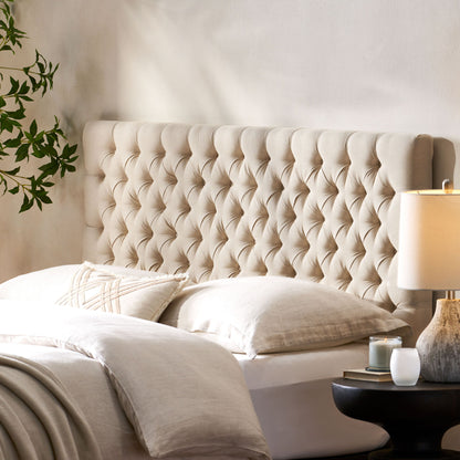 A cozy bedroom featuring the Annecy Contemporary Upholstered Headboard in beige, cream bedding, a round black nightstand with a lit candle and lamp, plus a leafy plant against a light-colored wall.