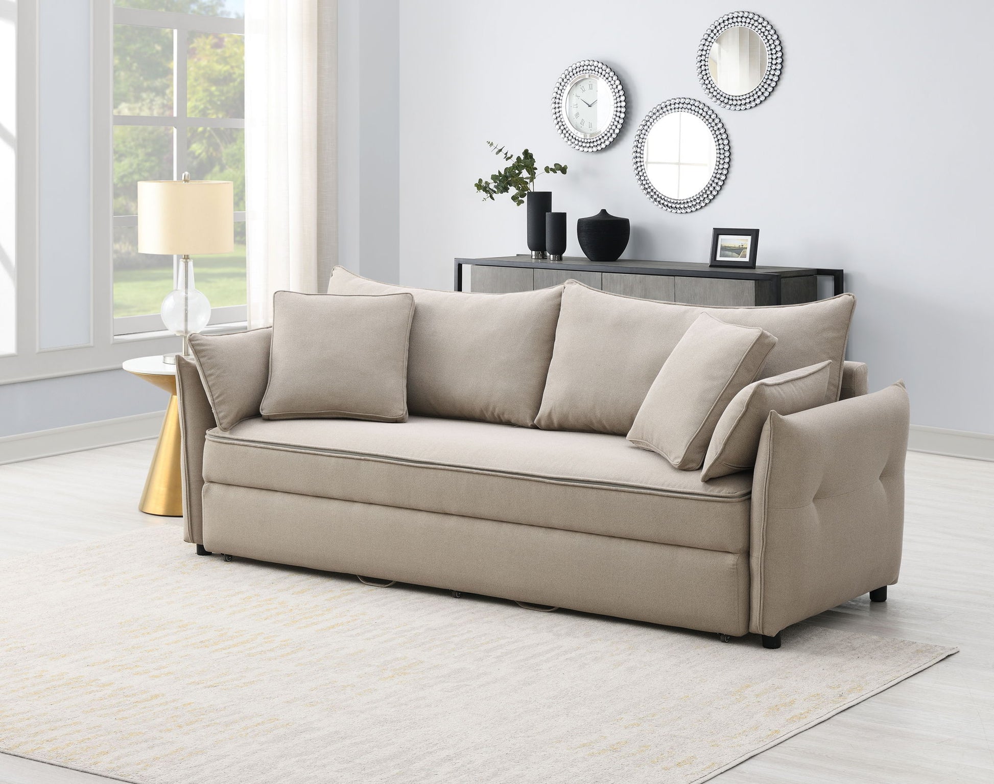 A modern living room with the Irina Pull Out Sleep With Storage beige sofa, matching pillows, a light area rug, a round gold side table with lamp, and a black console table topped with decorative mirrors and vases against a light wall.