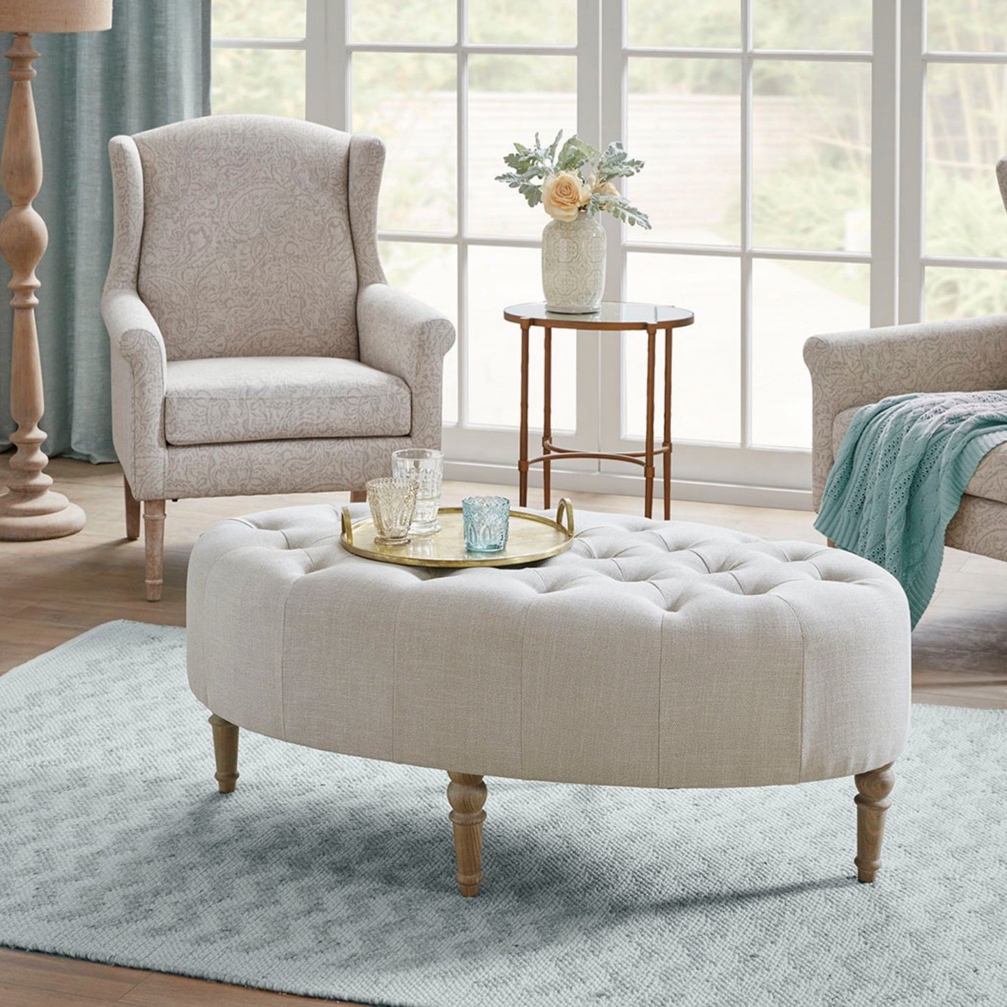 A cozy living room featuring the Fancy Style Design Upholstery Solid Wood - Linen armchair, a round ottoman, a serving tray with glassware, a side table with flowers, all on a light blue rug by large windows.