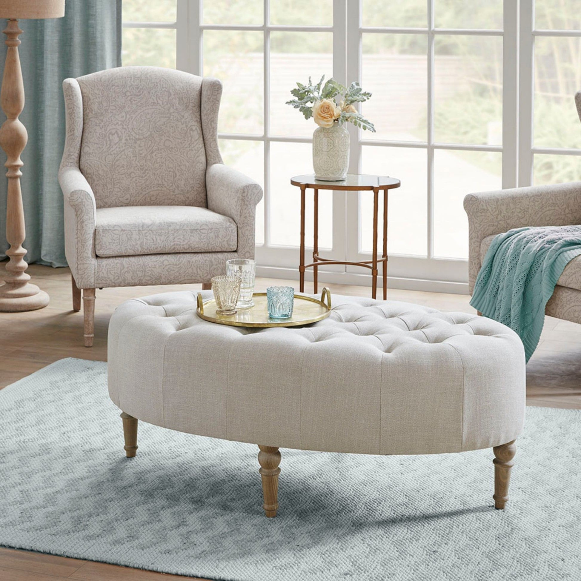 A cozy living room featuring the Fancy Style Design Upholstery Solid Wood - Linen armchair, a round ottoman, a serving tray with glassware, a side table with flowers, all on a light blue rug by large windows.