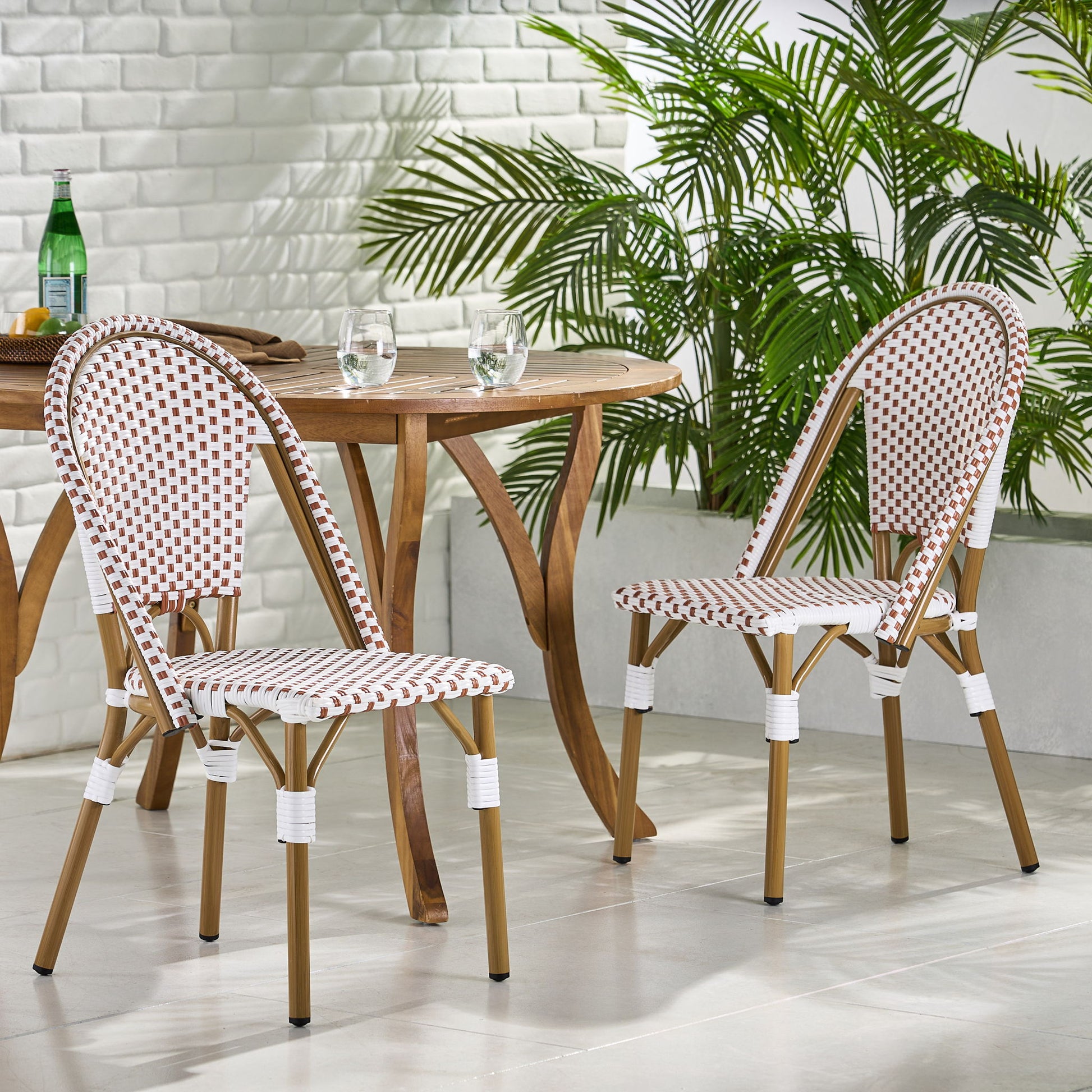 The Outdoor Rattan And Aluminum French Bistro Chairs Set, featuring red and white checkered seats and backs, is paired with a round wooden table holding two glasses and a bottle, surrounded by green plants and white brick walls on a modern patio.