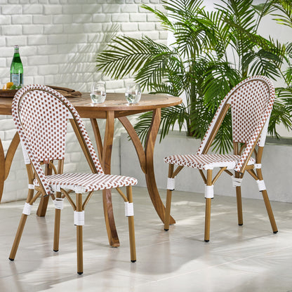 The Outdoor Rattan And Aluminum French Bistro Chairs Set, featuring red and white checkered seats and backs, is paired with a round wooden table holding two glasses and a bottle, surrounded by green plants and white brick walls on a modern patio.