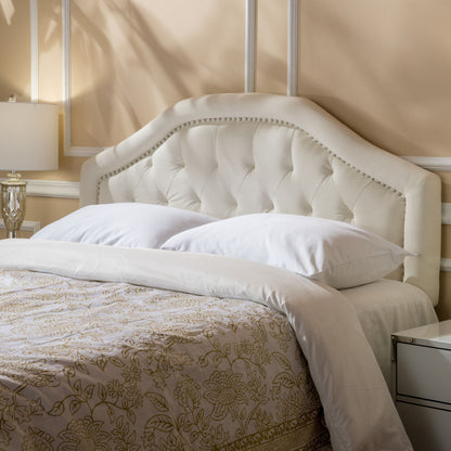 A neatly made bed featuring the Kaarina Contemporary Tufted Fabric Headboard in cream, two white pillows, a white comforter, and a beige floral blanket sits beside a white nightstand and lamp in a softly lit bedroom.