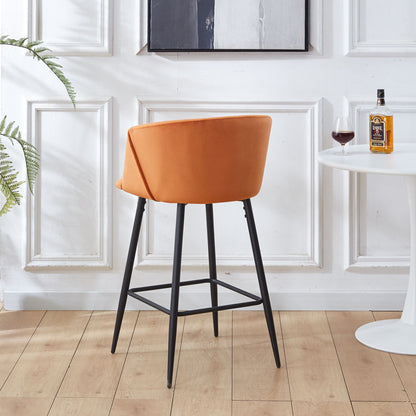 A pair of Counter Height Bar Stools with orange upholstery and black metal legs stand by a white round table holding a bottle, glass, and plant, against a white-paneled wall with framed art in a bright room with wood flooring.