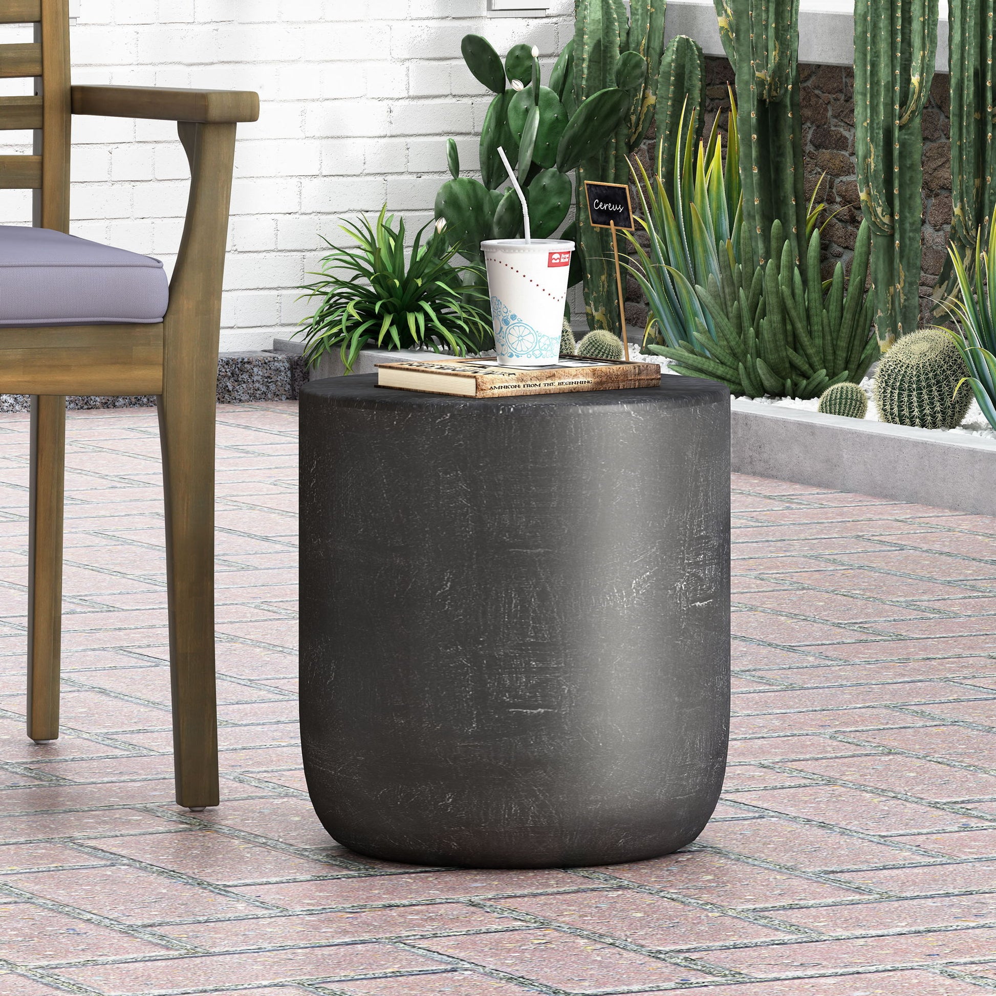 The Outdoor Lightweight Concrete End Table With Natural Texture - Matte Black sits on a tiled patio with a drink cup and book on top, near a wooden chair, green plants, and cacti by a white brick wall.