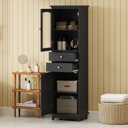 A tall black bathroom storage cabinet with glass doors, two drawers, and adjustable shelves displays towels and toiletries next to a small wooden shelf with mirrors and folded towels on a beige paneled wall.