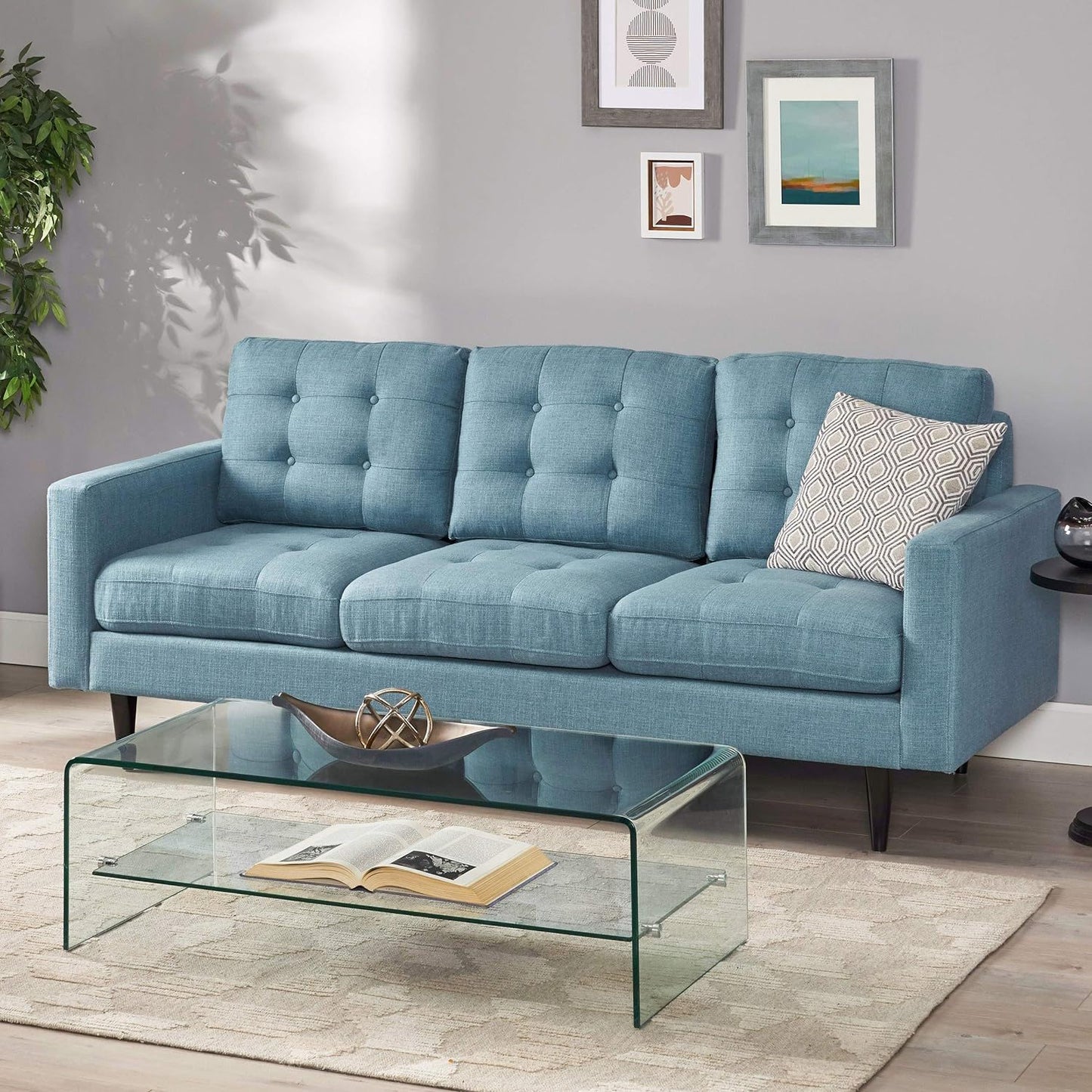 A modern living room with the Jenny contemporary tufted fabric 3 seater sofa in light blue, a geometric cushion, glass coffee table holding an open book and decorative bowl, beige rug, wall art, and a green potted plant in the corner.