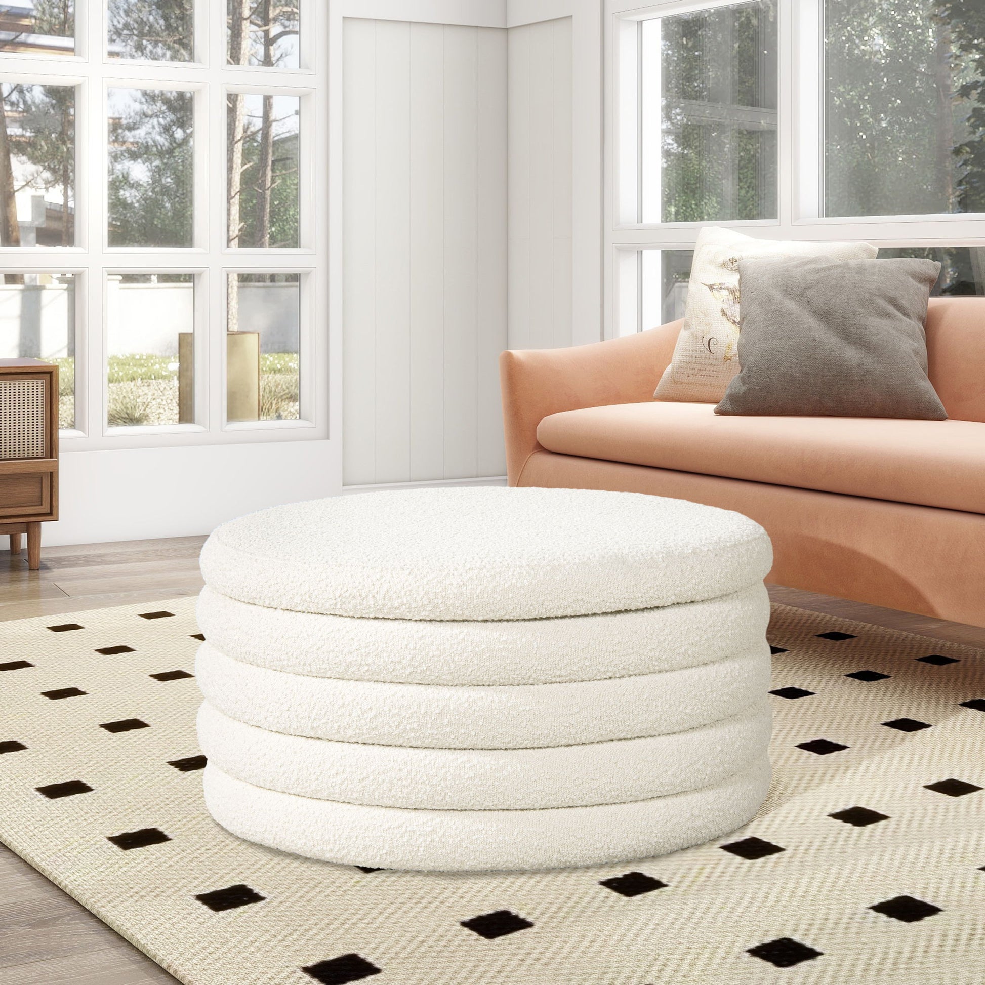 A modern living room with a large window, tan sofa with throw pillows, and the Fuji Oversized Round Storage Ottoman in white on a beige rug featuring black square patterns.