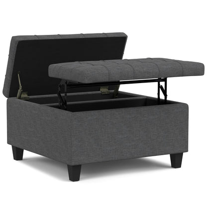 The Harrison Upholstered Coffee Table Storage Ottoman features a gray tufted, lift-top lid that opens to reveal spacious interior storage and stands on black wooden legs.