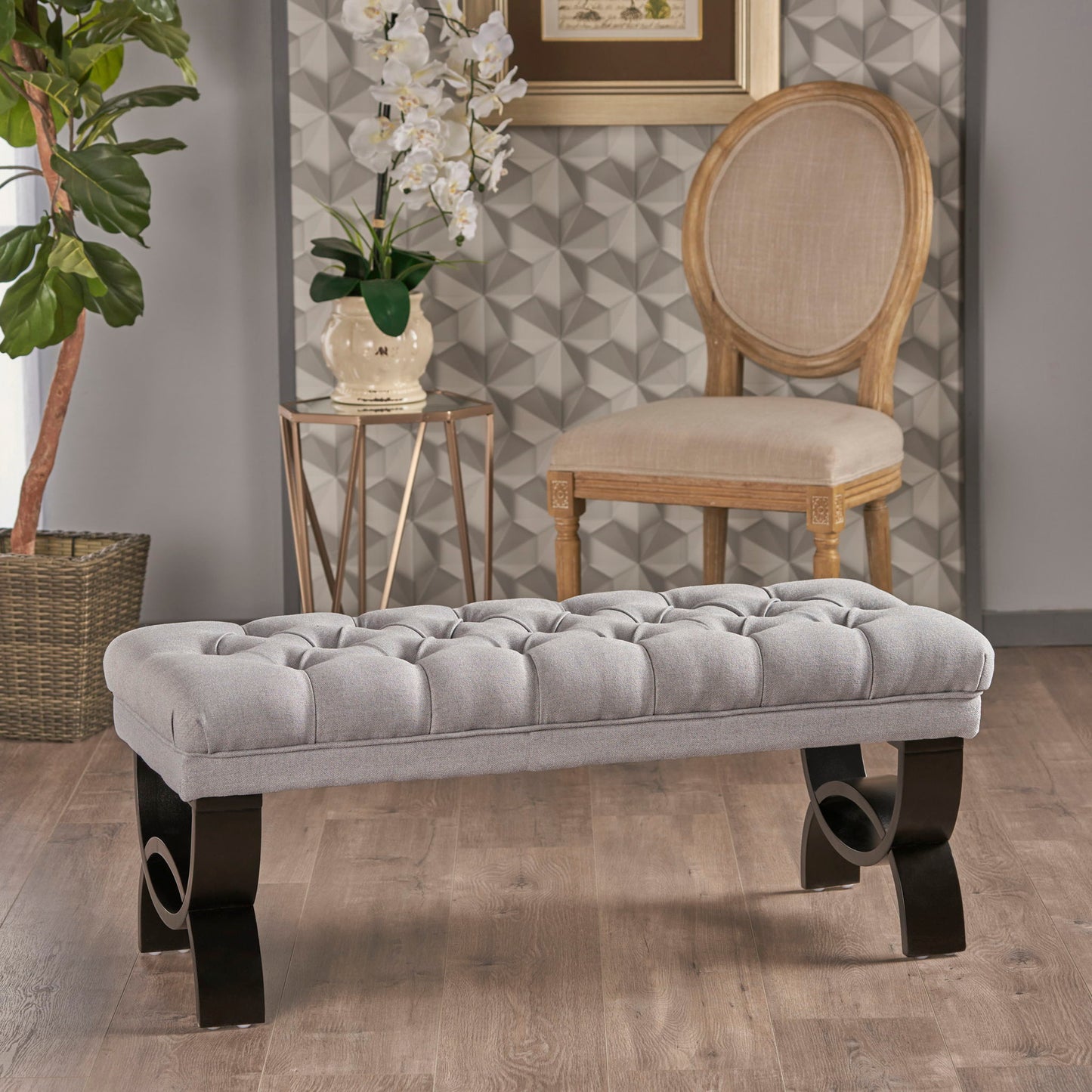 The Hilton - Ottoman Bench Classic Style, featuring tufted light gray upholstery and dark curved legs, rests on a wooden floor. Behind it are a beige chair, geometric-patterned wall, potted plant, table with orchids, and framed picture.