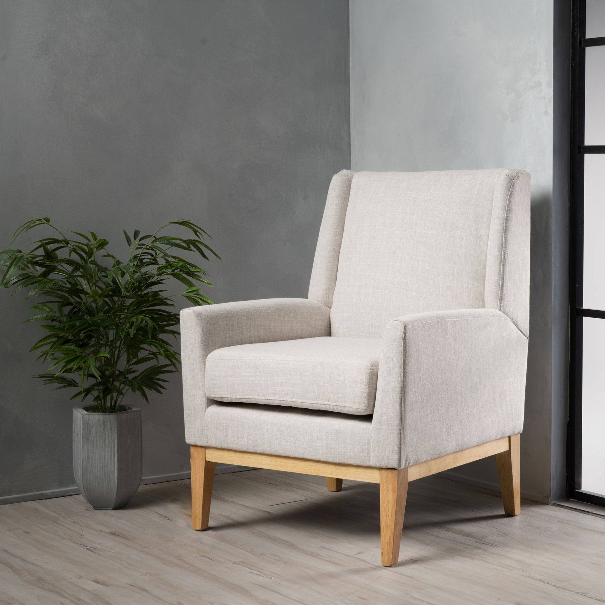 The Modern Accent Chair With Padded Cushion Seat, featuring beige upholstery and wooden legs, is placed in a modern room with light wood floors, gray textured walls, and a potted green plant in the corner.