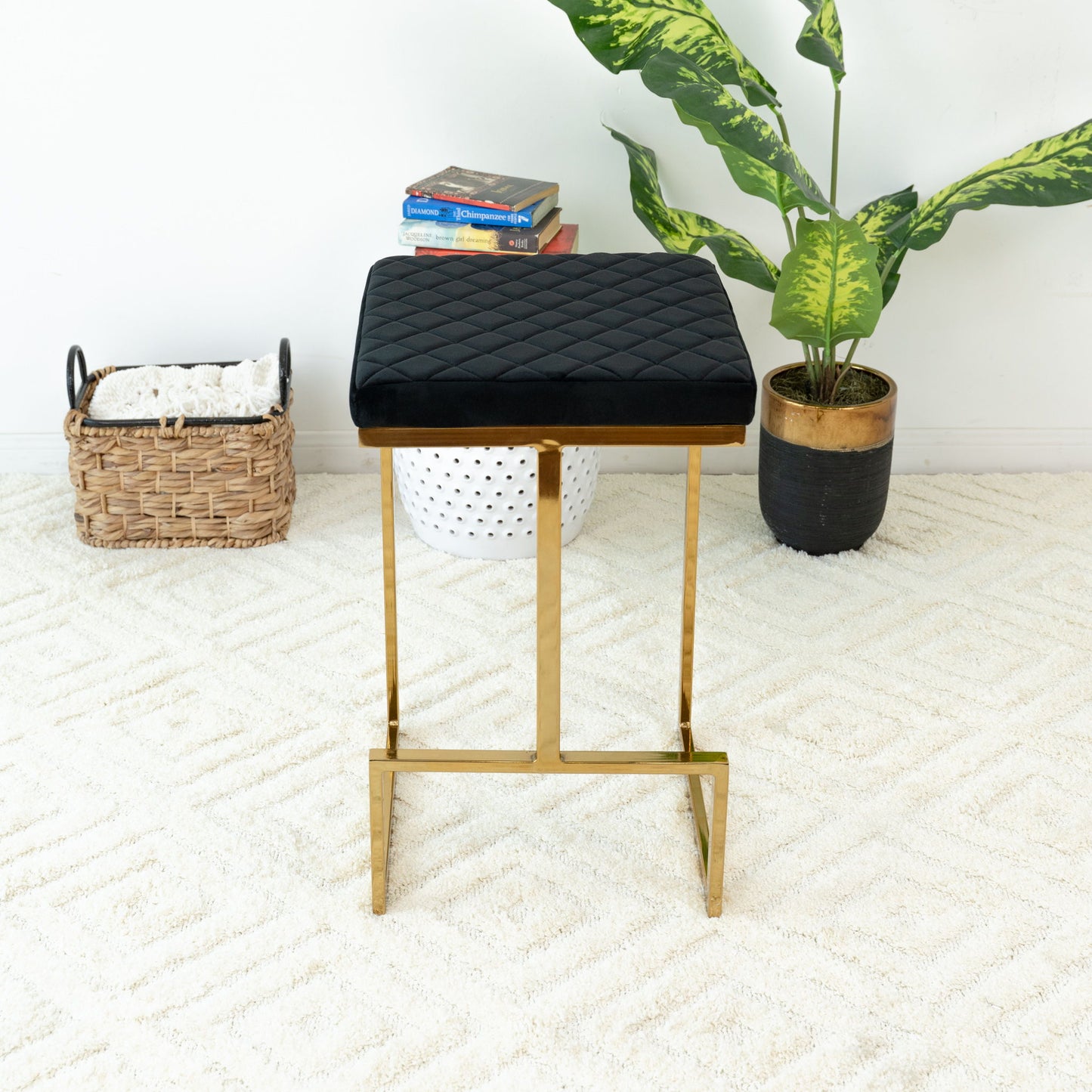 The Joel - Mid Century Modern Luxury Upholstered Stool, featuring a black cushion and gold metal legs, stands on a white textured rug beside a potted plant, woven basket with white blanket, white basket, and books.