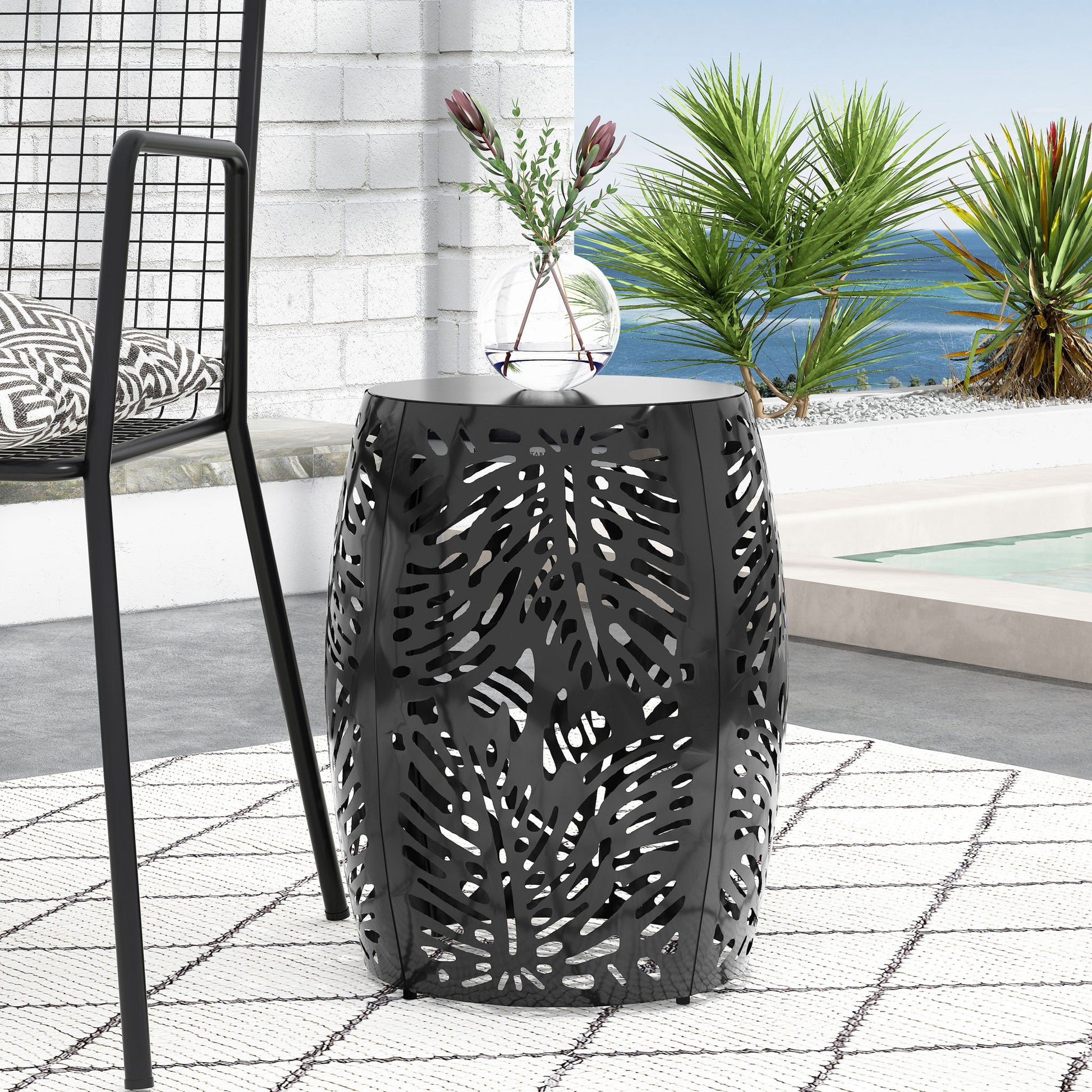 The Modern Design Metal End Table, Large, with a black cutout leaf pattern, sits on a patterned rug next to a modern chair. Tropical plants, a pool, and an ocean view under a clear sky fill the background.