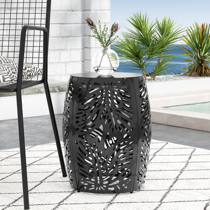 The Modern Design Metal End Table, Large, with a black cutout leaf pattern, sits on a patterned rug next to a modern chair. Tropical plants, a pool, and an ocean view under a clear sky fill the background.
