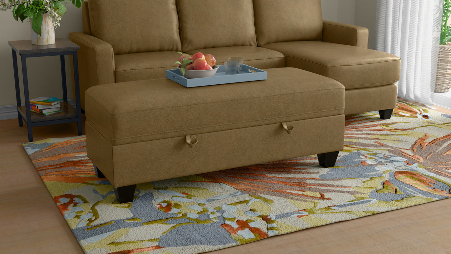 The Drason Storage Ottoman, upholstered in tan, sits on a colorful floral rug before a matching sectional. A blue tray with apples and glasses rests atop, while sunlight pours through a window dressed in white curtains.