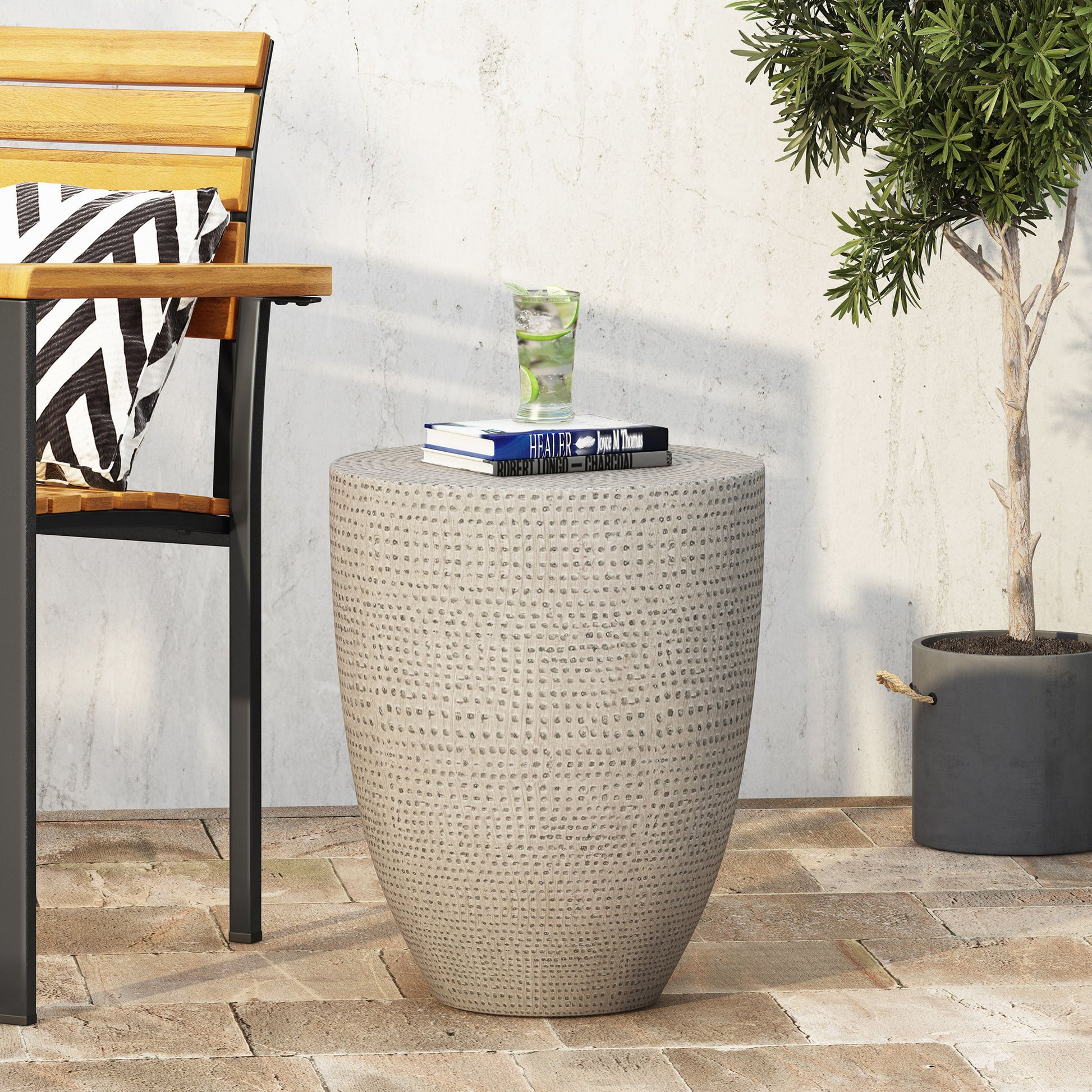 The Outdoor Lightweight Concrete Side Table in Natural features a textured beige surface and compact round design, perfect for patios. Pair it with a black metal-frame wooden chair and striped cushion beside a potted plant for a cozy setting.