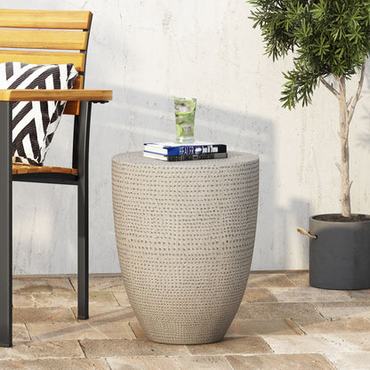 The Outdoor Lightweight Concrete Side Table in Natural features a textured beige surface and compact round design, perfect for patios. Pair it with a black metal-frame wooden chair and striped cushion beside a potted plant for a cozy setting.