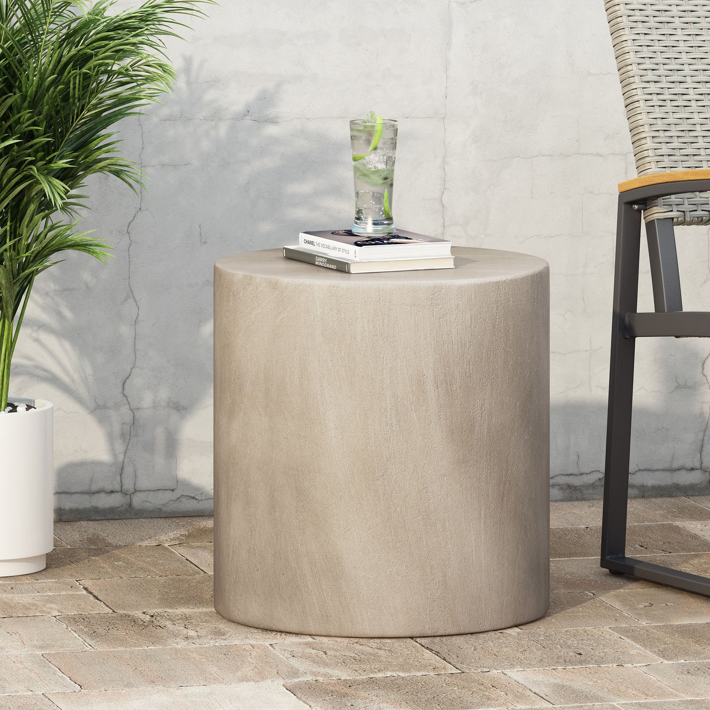 A Cylindrical Design, Outdoor Lightweight Concrete Side Table holds a glass of lime water and books. It sits on a brick patio next to a chair and potted plant, set against a light-colored wall.