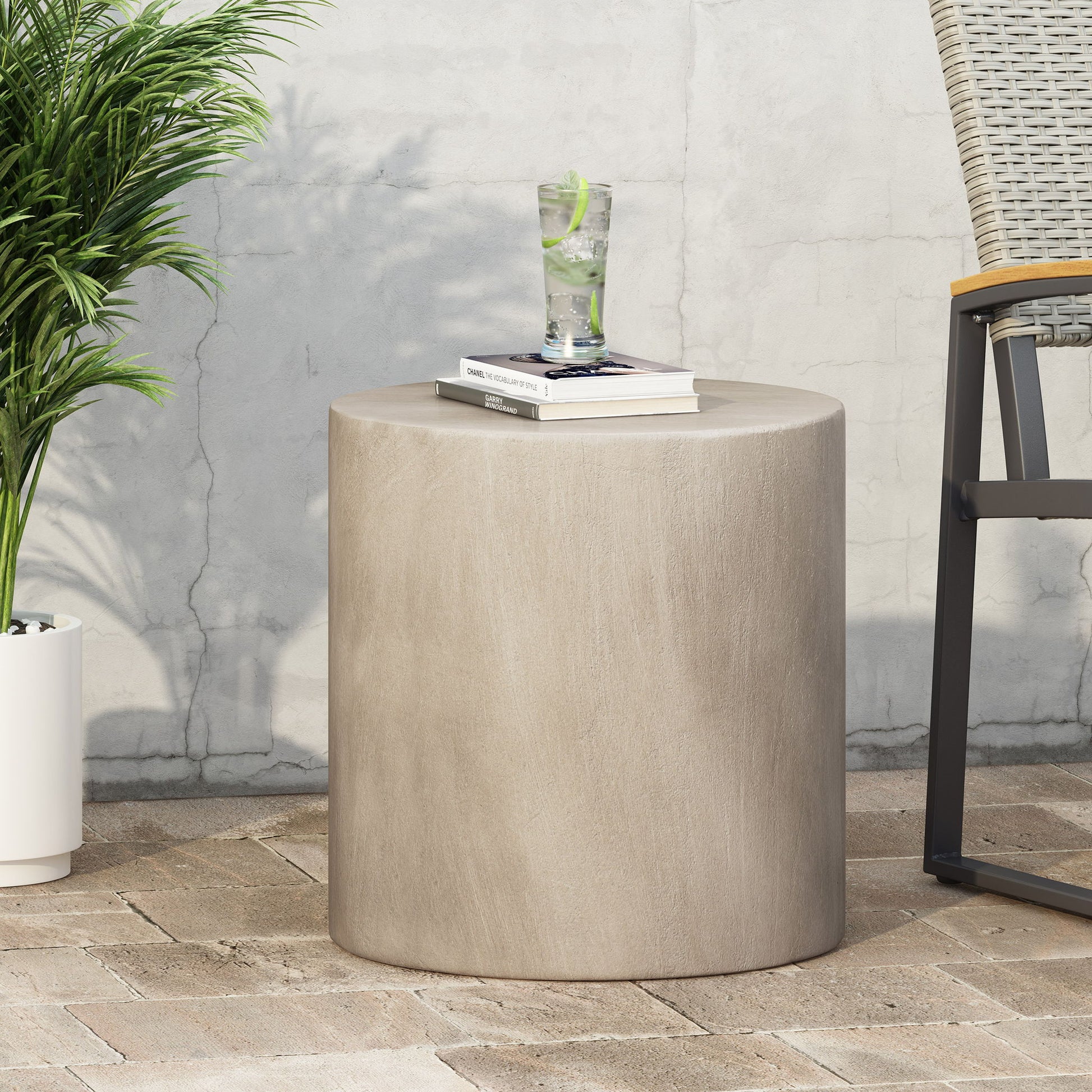 A Cylindrical Design, Outdoor Lightweight Concrete Side Table holds a glass of lime water and books. It sits on a brick patio next to a chair and potted plant, set against a light-colored wall.