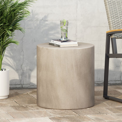 A Cylindrical Design, Outdoor Lightweight Concrete Side Table holds a glass of lime water and books. It sits on a brick patio next to a chair and potted plant, set against a light-colored wall.