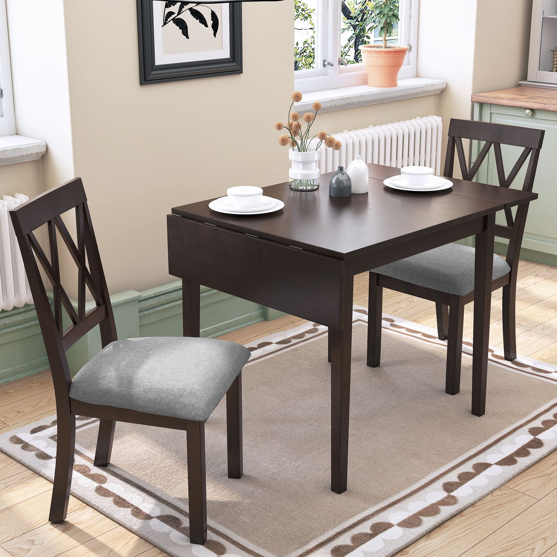 The 3 Piece Kitchen Dining Set features a drop-leaf table and two upholstered chairs, perfect for small spaces. Styled with white plates and dried flowers, the set sits on a beige rug in a bright room with window and framed artwork.