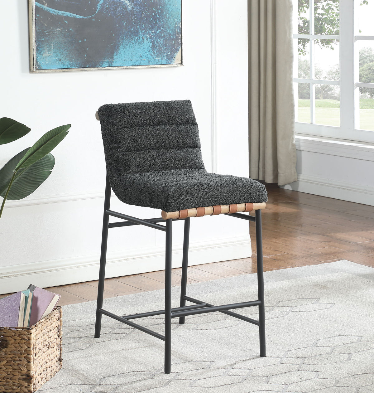 The Lahni 17" Boucle Fabric Counter Height Chair features a black metal frame and a textured dark gray cushioned seat, shown on a light rug in a bright room with wooden floors, large windows, a plant, and wall art.