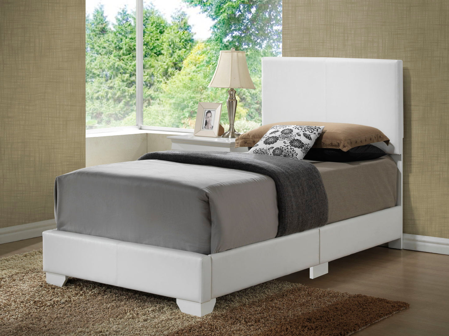 The Elegant Platform Bed For Relaxed Spaces features a white frame and headboard, gray and brown bedding, decorative pillows, and sits beside a large window with green views, complemented by a bedside table with lamp and picture frame.