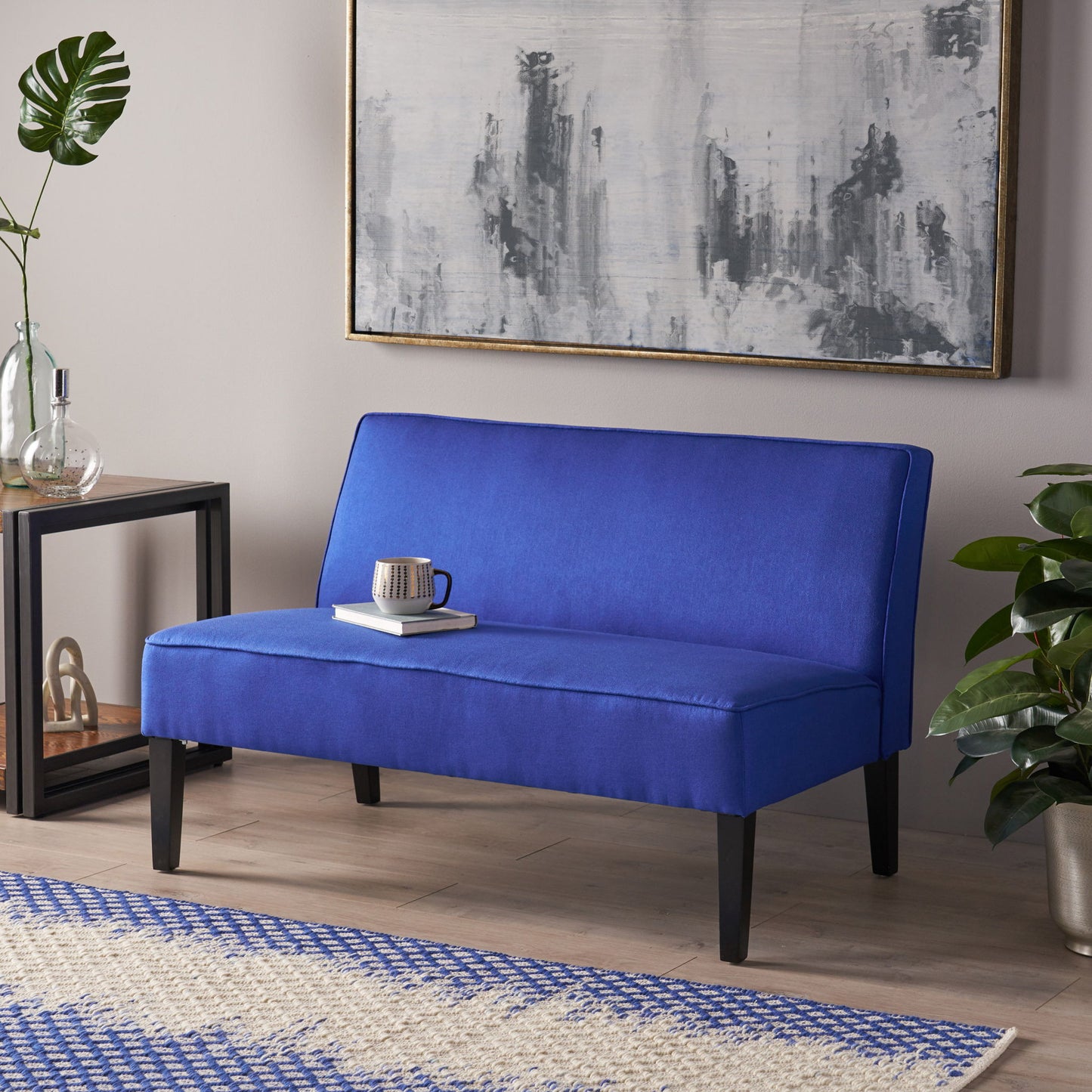A contemporary blue upholstered loveseat with birch wood legs sits against the wall, topped with a cup and book. Abstract art hangs above, a decorated side table is on the left, and a plant is positioned on the right.
