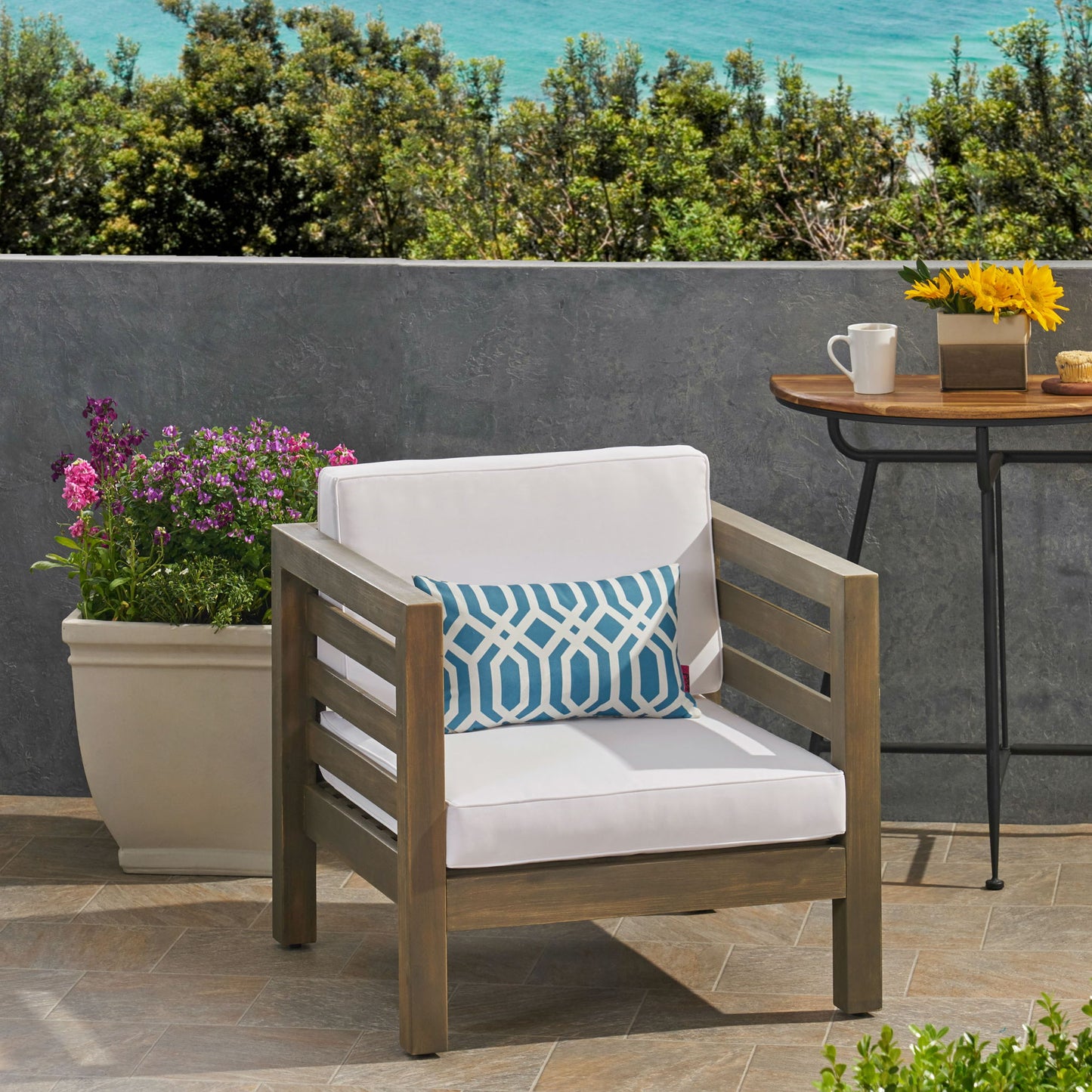 An Outdoor Acacia Wood Patio Chair With Fabric Cushion, complemented by a blue patterned pillow, is on a tiled terrace beside a large potted plant and a table with coffee, sunflowers, and an ocean view in the background.