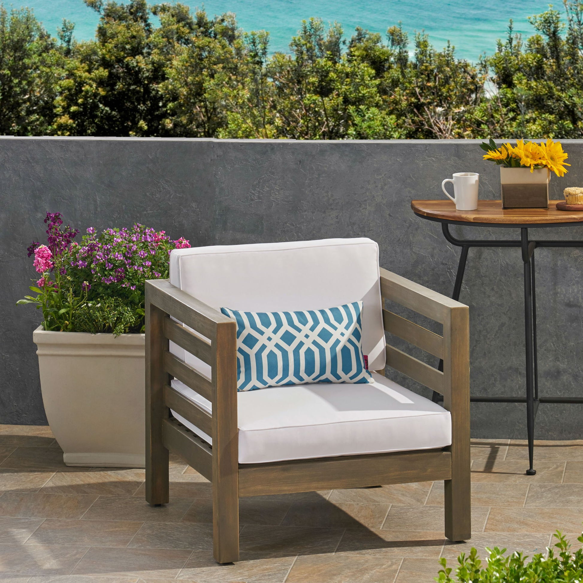 An Outdoor Acacia Wood Patio Chair With Fabric Cushion, complemented by a blue patterned pillow, is on a tiled terrace beside a large potted plant and a table with coffee, sunflowers, and an ocean view in the background.