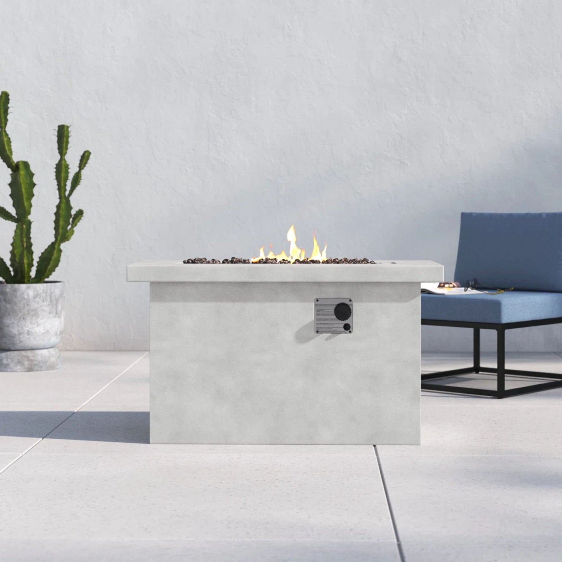 A Propane / Gas Outdoor Fire Pit Table with visible flames stands on a light patio. In the background, there’s a potted cactus and a blue cushioned black metal chair set against a white wall.