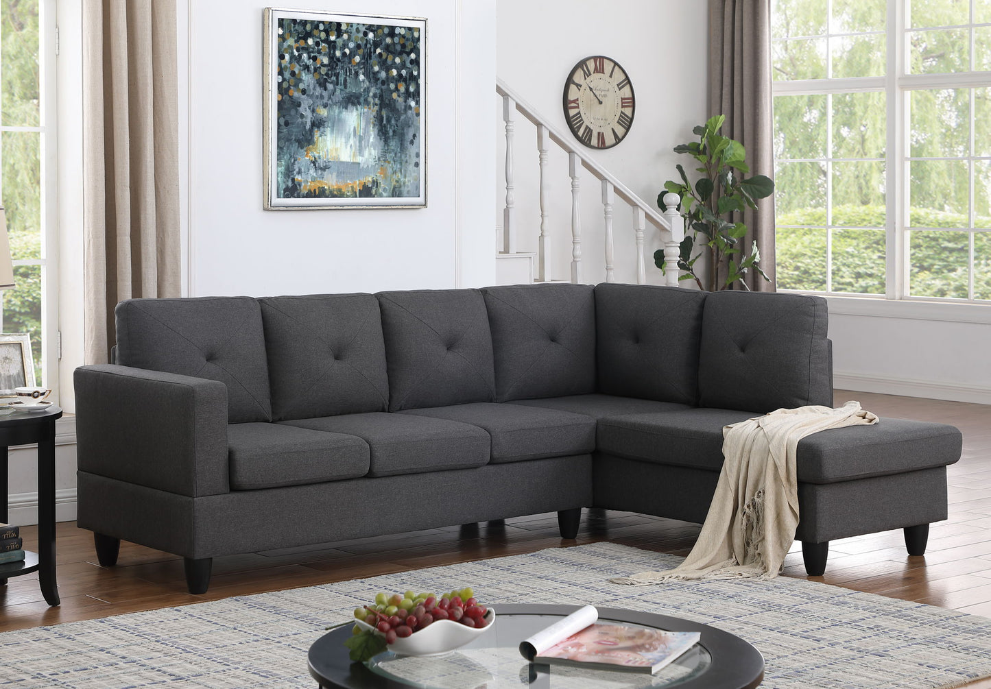 A modern living room with the Santiago Linen Sectional Sofa with right facing chaise in dark gray, a round coffee table holding fruit and books, a wall clock, leafy plant, large windows with light curtains, and a framed abstract painting.