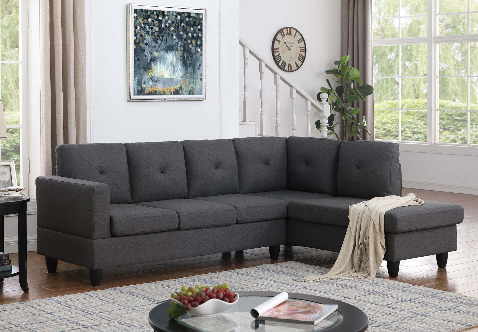 A modern living room with the Santiago Linen Sectional Sofa with right facing chaise in dark gray, a round coffee table holding fruit and books, a wall clock, leafy plant, large windows with light curtains, and a framed abstract painting.