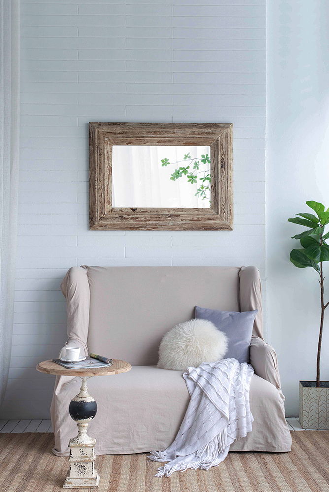 A cozy living room features a beige loveseat with a white round pillow, gray cushion, and white throw. A rustic side table holds a teacup. A plant and the Rectangle Wall Accent Mirror With Distressed Wood Frame - Brown add charm.