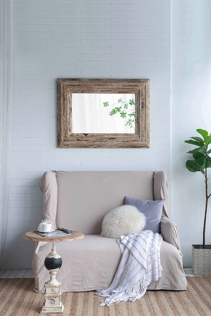 A cozy living room features a beige loveseat with a white round pillow, gray cushion, and white throw. A rustic side table holds a teacup. A plant and the Rectangle Wall Accent Mirror With Distressed Wood Frame - Brown add charm.