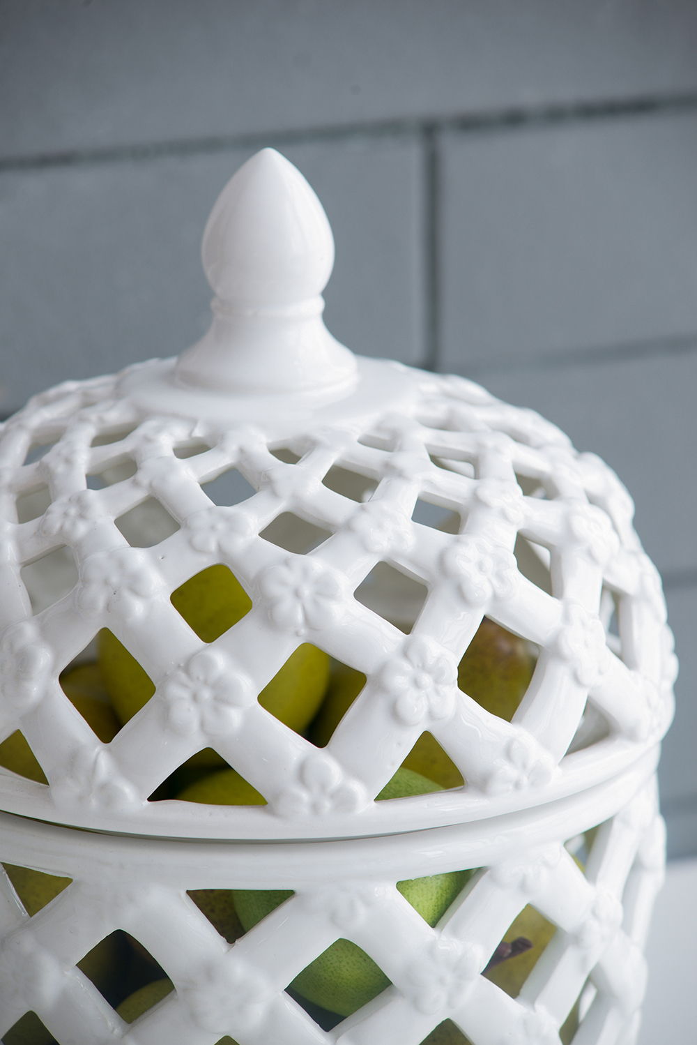 A close-up of the Ballas - Bird Lidded Jar in white, a ceramic piece with a diamond lattice pattern and floral accents; green pears are visible inside through the openwork design.