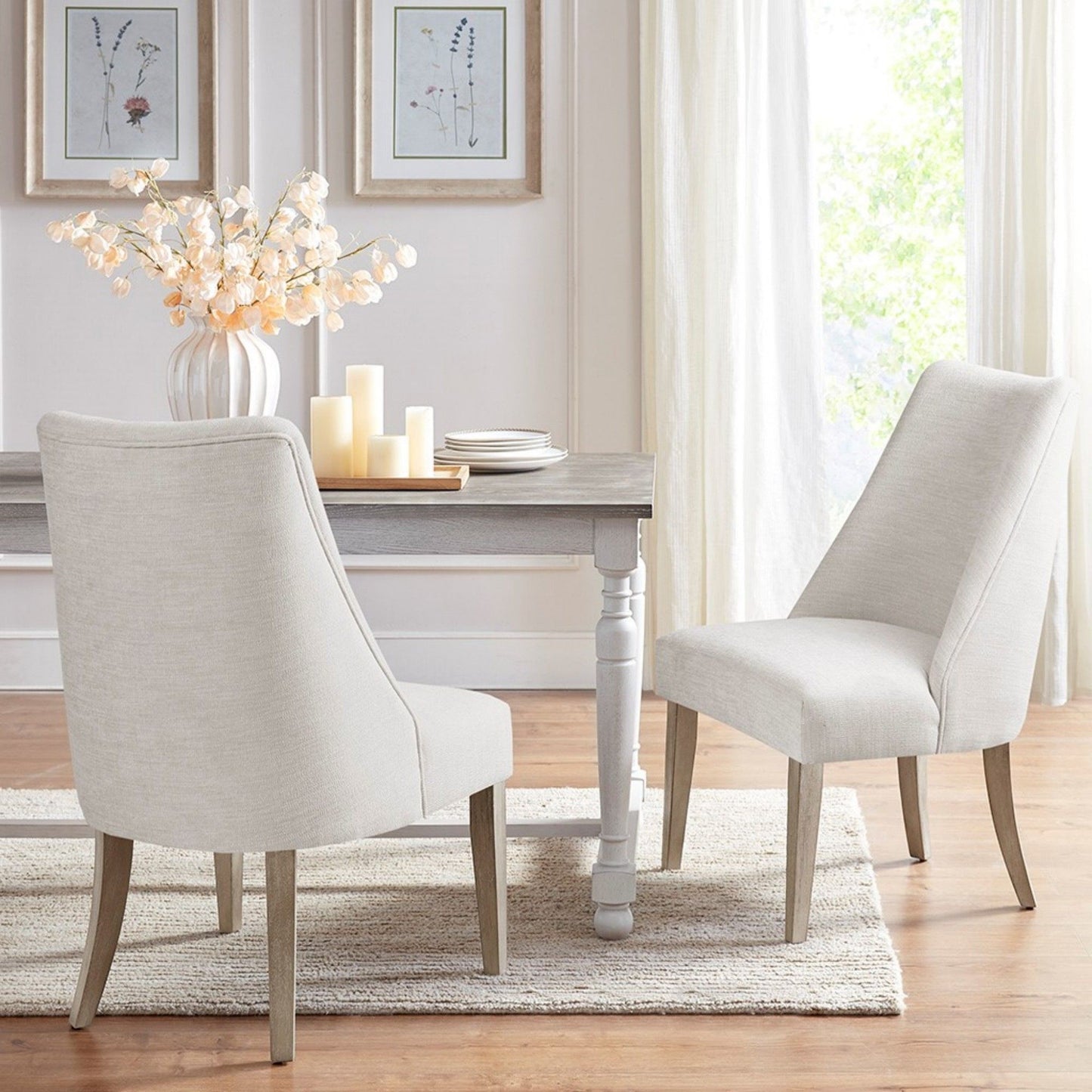 Two Comfortable Dining Chair Upholstered (Set of 2), in cream, are placed at a wooden dining table decorated with white flowers and candles. The space features light walls, large windows with curtains, framed art, wood floors, and a beige rug.