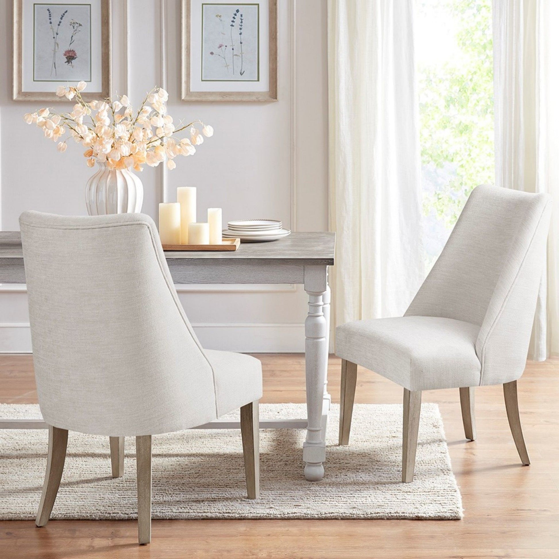 Two Comfortable Dining Chair Upholstered (Set of 2), in cream, are placed at a wooden dining table decorated with white flowers and candles. The space features light walls, large windows with curtains, framed art, wood floors, and a beige rug.