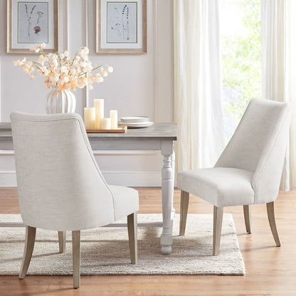 Two Comfortable Dining Chair Upholstered (Set of 2), in cream, are placed at a wooden dining table decorated with white flowers and candles. The space features light walls, large windows with curtains, framed art, wood floors, and a beige rug.