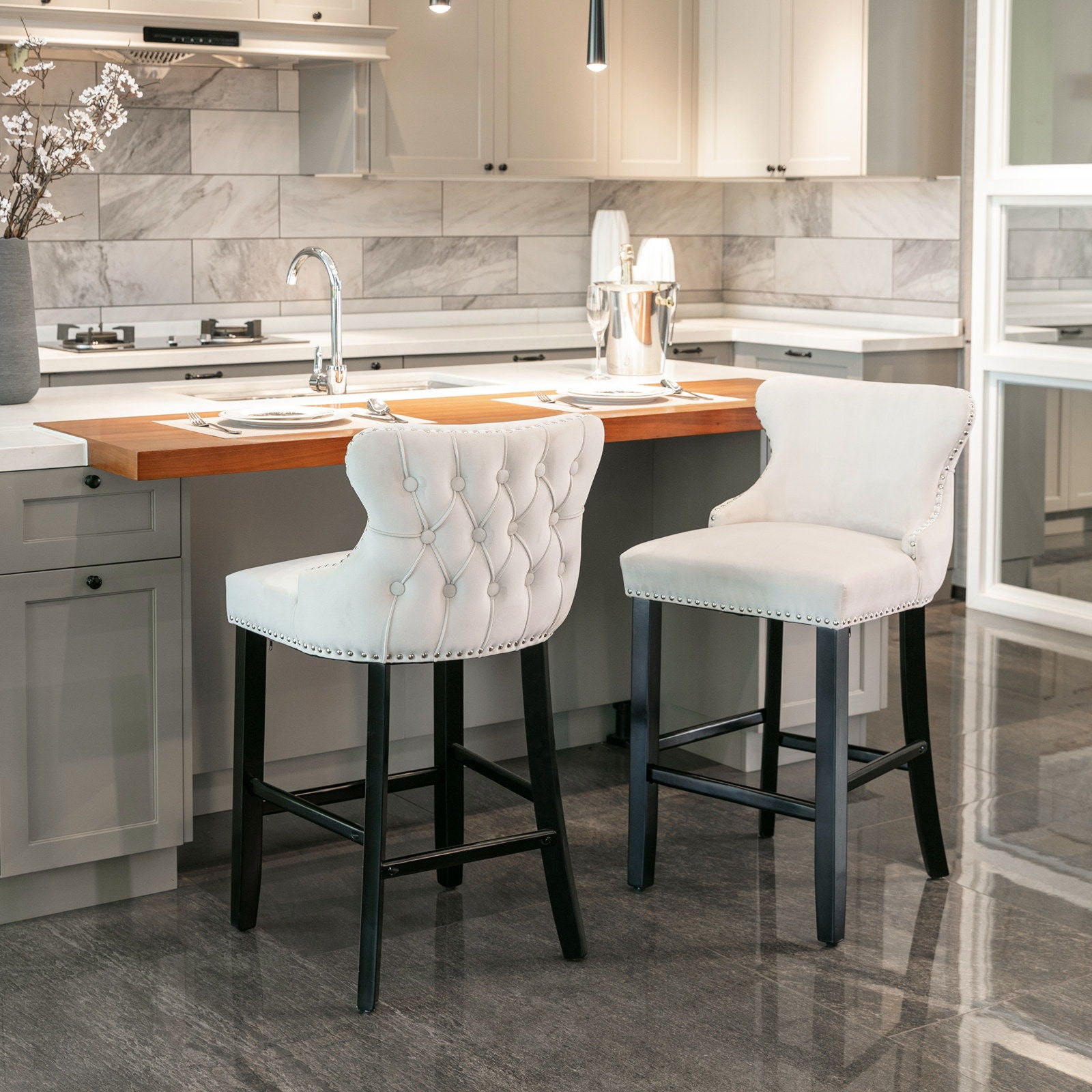A modern kitchen with a marble backsplash, light gray cabinets, and a wooden breakfast bar featuring two Contemporary Velvet Wing-Back Barstools with button tufting, chrome nailhead trim, and dark wooden legs. Table settings and a vase are displayed.