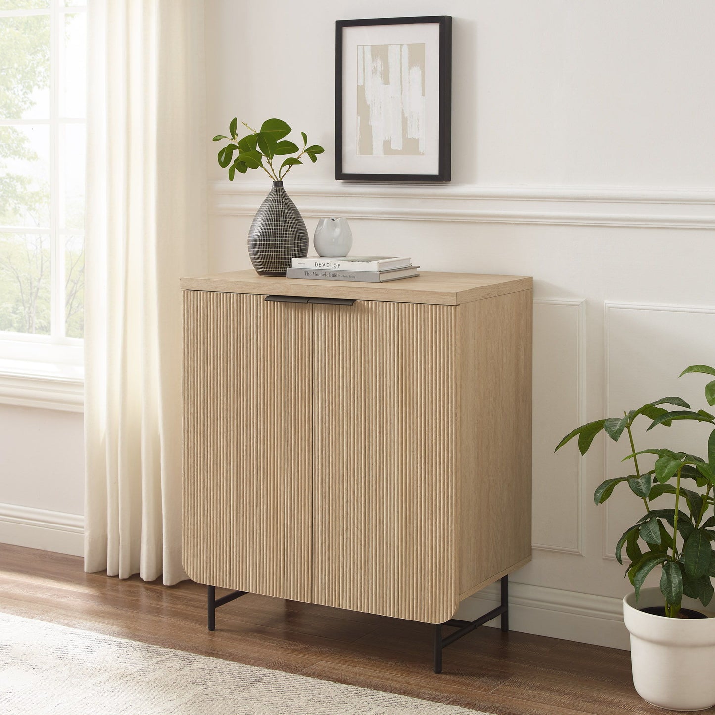 The Couch.com Modern Scandi Fluted Door Storage Console in coastal oak stands on black legs by a window with white curtains. On top are a plant, gray vase with greenery, books, and above hangs abstract art. A potted plant sits nearby.