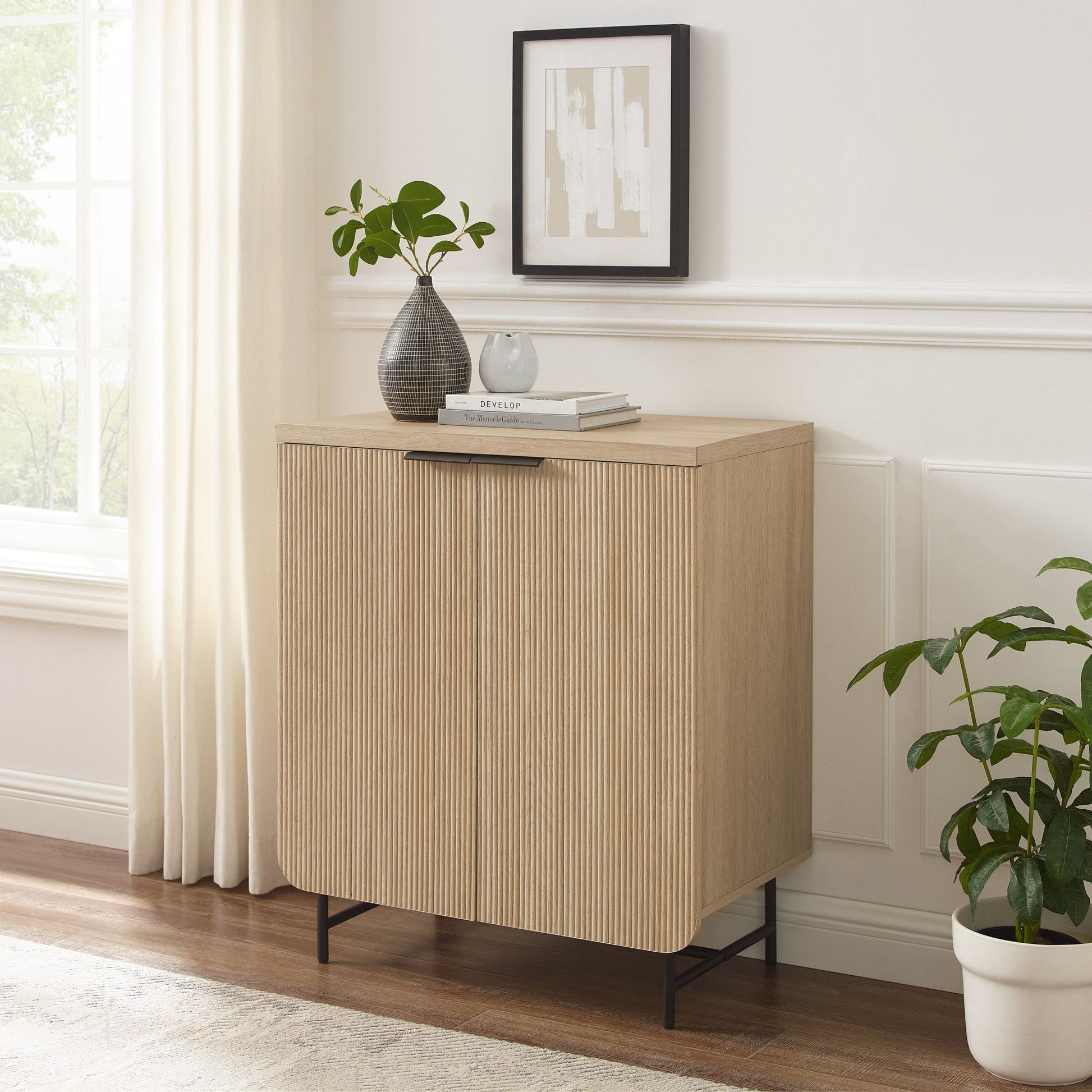The Couch.com Modern Scandi Fluted Door Storage Console in coastal oak stands on black legs by a window with white curtains. On top are a plant, gray vase with greenery, books, and above hangs abstract art. A potted plant sits nearby.