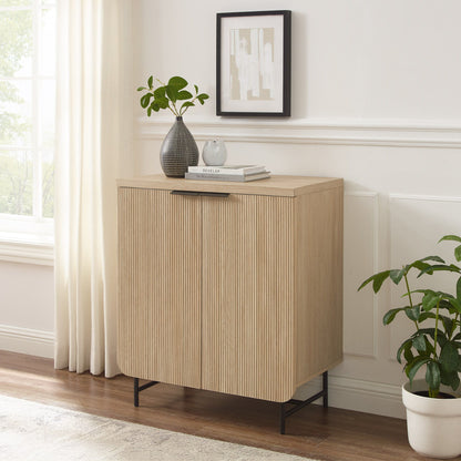 The Couch.com Modern Scandi Fluted Door Storage Console in coastal oak stands on black legs by a window with white curtains. On top are a plant, gray vase with greenery, books, and above hangs abstract art. A potted plant sits nearby.