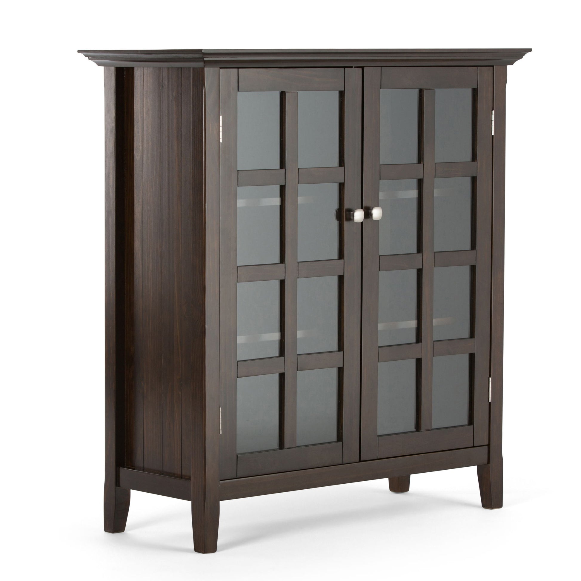 The Acadian - Medium Storage Cabinet is handcrafted from dark wood, featuring two grid-patterned glass doors with metal knobs, four sturdy legs, and a slightly overhanging top.