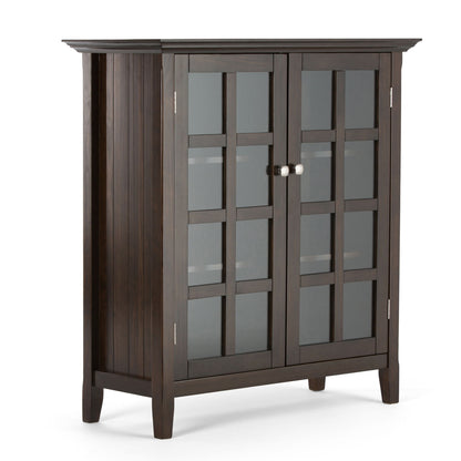 The Acadian - Medium Storage Cabinet is handcrafted from dark wood, featuring two grid-patterned glass doors with metal knobs, four sturdy legs, and a slightly overhanging top.