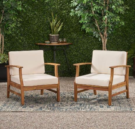Two Perla Contemporary Club Chairs with water-resistant cushions are placed on a patterned rug over gravel, with a small table, plants, and a hedge backdrop creating a cozy outdoor seating area.