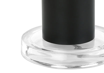 Close-up of the base of a contemporary table lamp in black, resting on a round, transparent glass or acrylic platform against a white background.