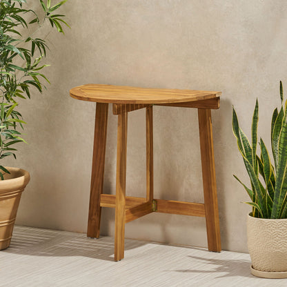 The Outdoor Half Round Folding Acacia Wood Bistro Table stands against a beige wall, flanked by green potted plants. It adds a minimal and natural touch with its angled legs and compact wooden design.