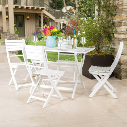 The Positano - Foldable Dining Set, featuring a white table and four chairs, is arranged on a light stone patio. The table is set with wine glasses, a wine bottle, flowers, and decor. Lush greenery and a house provide a scenic backdrop.