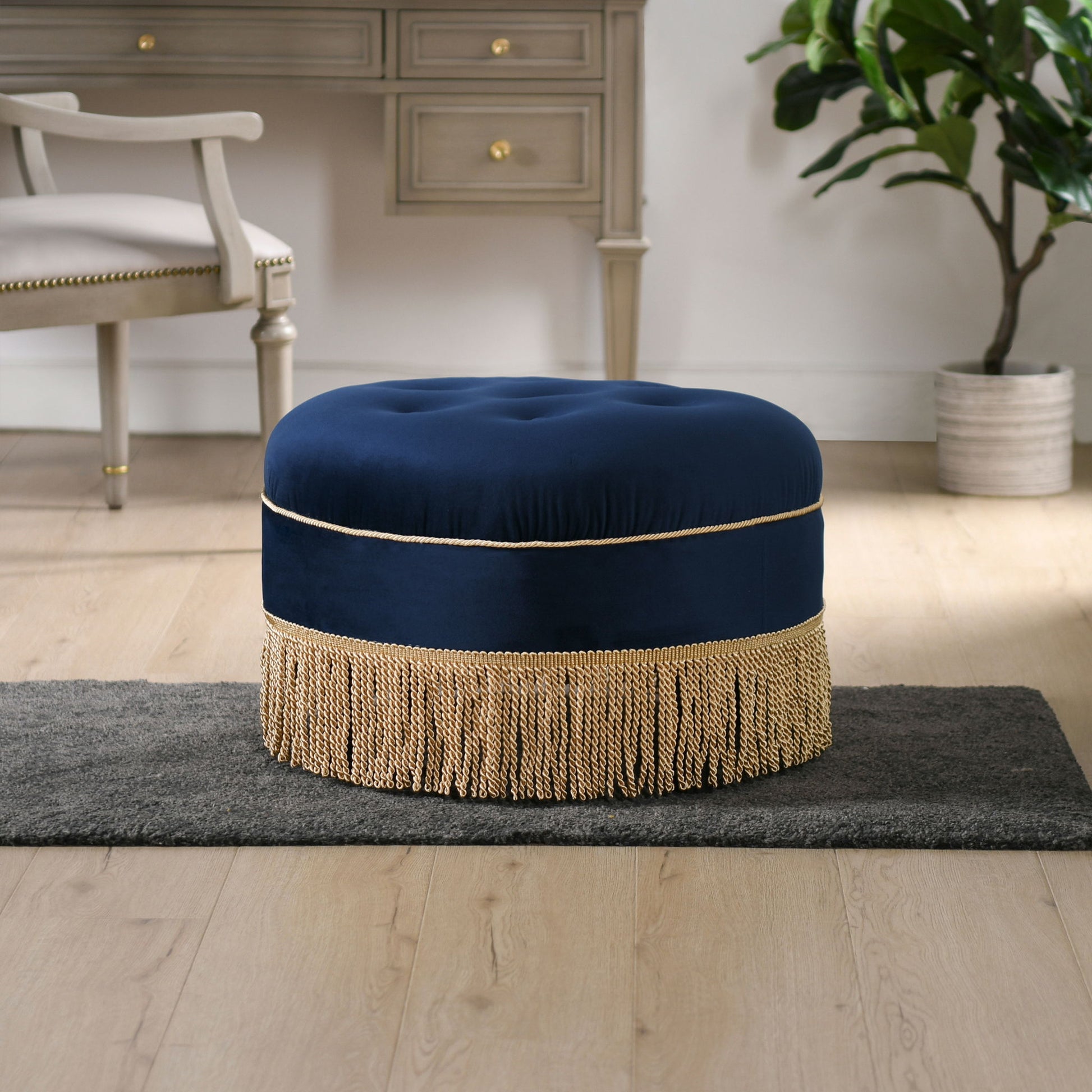 The Yolanda Round Upholstered Accent Ottoman, featuring navy blue velvet and gold fringe trim, rests on a gray rug in a light wood-floored room with a desk, chair, and potted plant in the background.